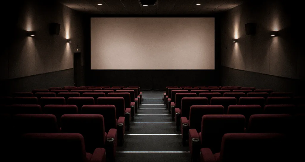 Salle de cinéma aux fauteuils rouges disposés en gradins face à un grand écran blanc, éclairée par une lumière tamisée.
