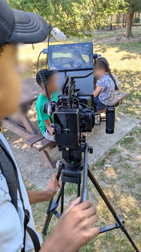 Camera rig on a tripod with external monitor person operating it outdoors while two children sit on a bench in a sunny park faces blurred