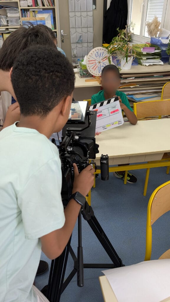 Two students in a classroom one operates a video camera on a tripod while the other holds a clapperboard for a filming project